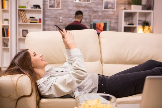 Beautiful young woman resting on sofa and browsing on sofa. Boyfriend in the background.
