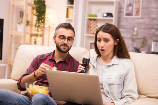 Caucasian couple drinking soda and eating chips while watching a movie on laptop. Couple sitting on couch.
