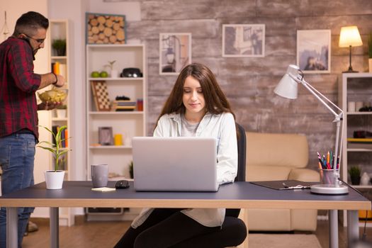 Girlfriend working on laptop in living room and boyfriend talking on the phone and eating chips in the background.