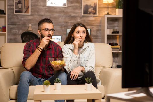 Couple eating chips and looking shocked at tv while watching a movie at night.
