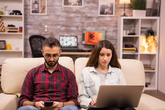 Caucasian beautiful couple with serious faces sitting on sofa. Couple using laptop and phone while sitting on sofa.