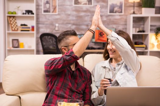 Couple giving high five while watching a movie on laptop and sitting on sofa. Chips and soda.