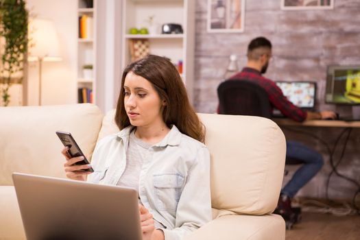 Beautiful young woman sitting on sofa with laptop and browsing on phone. Boyfriend in the background.