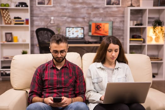 Caucasian beautiful couple with serious faces sitting on sofa. Couple using laptop and phone while sitting on sofa.