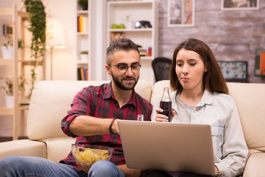 Caucasian couple drinking soda and eating chips while watching a movie on laptop. Couple sitting on couch.
