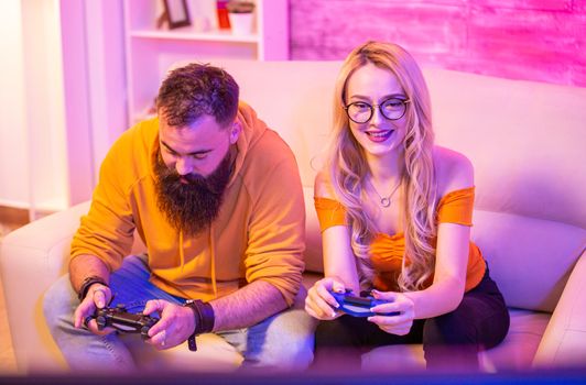 Cheerful young couple sitting on sofa and playing video games using wireless controllers. Room with colorful neon light.