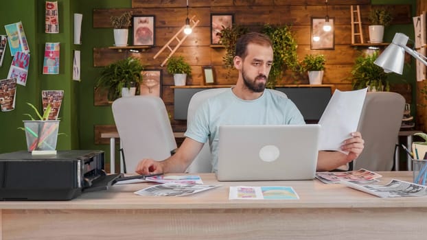 Young designer looking at a sheet holding it in his hand while sitting at the table in front of a laptop. Caucasian creativ artist