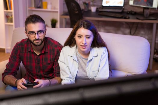 Joyful young couple playing video games on television using controllers at night. Couple sitting on couch.