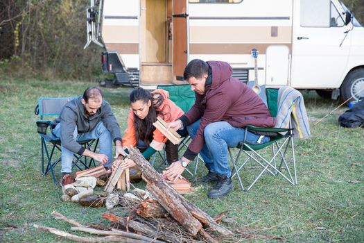Group of close friends setting up the camp fire in the woods with retro camper van in the background. Camping in mountains.