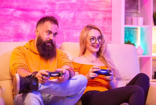 Beautiful blonde girl smiling while playing video games with her boyfriend using wireless controllers on a big screen TV