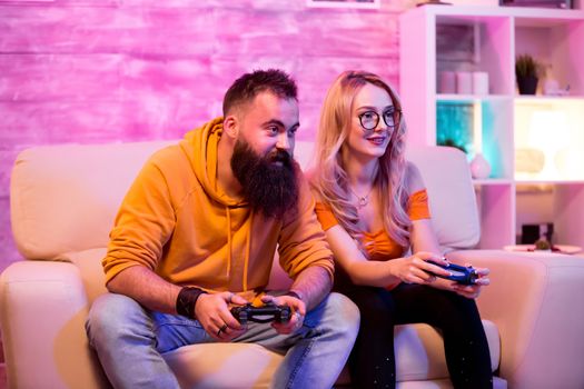 Young man and woman using wireless controller to play online games sitting on sofa. Room with neon light.