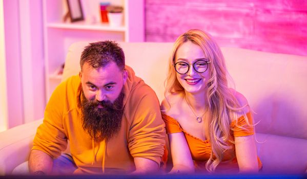 Beautiful blonde girl smiling while playing video games with her boyfriend using wireless controllers on a big screen TV