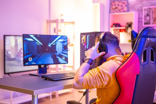 Bearded man playing online video games on his pc and talking with others players. Colorful neons in the room.