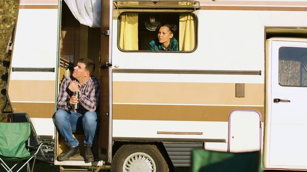Boyfriend sitting on the stairs of camper van. Girlfriend admiring the wild scenery from the window of camper van. Camping tent.