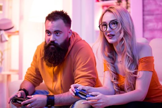 Couple sitting on sofa and playing online video games using wireless controler. Room with neon light.