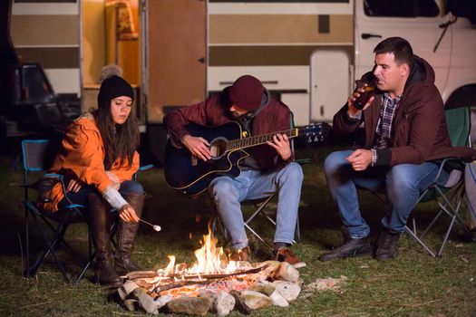 Tourists in a campsite playing on guitar and roasting marshmallows. Retro camper van. Camping in mountanis.