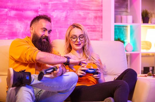 Beautiful blonde girl smiling while playing video games with her boyfriend using wireless controllers on a big screen TV