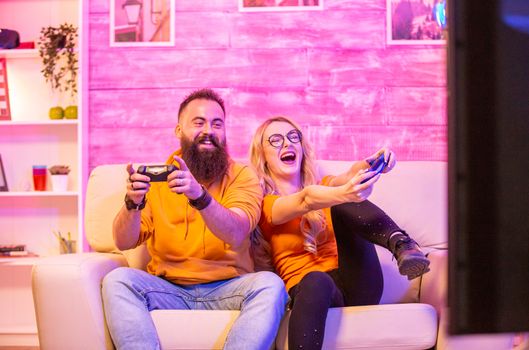 Beautiful blonde girl smiling while playing video games with her boyfriend using wireless controllers on a big screen TV