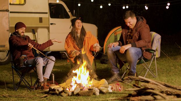 Bearded man telling a funny joke to his friends around camp fire. Retro camper van. Camping tent.