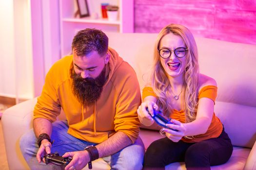 Cheerful young couple sitting on sofa and playing video games using wireless controllers. Room with colorful neon light.
