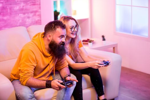 Bearded boyfriend helping his blond girlfriend to win at online games using wireless controller. Room with neon light.