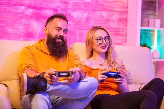 Beautiful blonde girl smiling while playing video games with her boyfriend using wireless controllers on a big screen TV