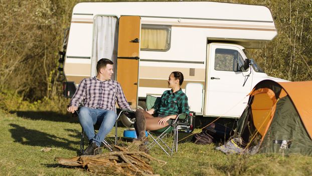 Hipster boyfriend kissing his girlfriend hand while sitting on camping seats in front of their vintage camper van. Relaxing Week-end