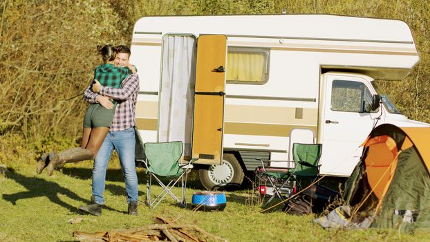 Boyfriend spinning around her girlfriend in front of their retro camper van. Couple with vintage camper in wilderness. Camping tent.