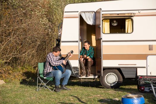 Man playing playing some good music on guitar to his girlfriend in front of their retro camper van.