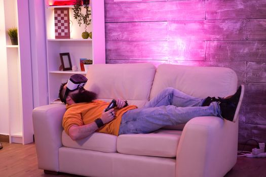 Male sitting on couch with virtual reality goggles in a room with colorful neon light.