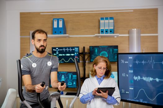 Male athlete who makes a physical effort on the stepper and has electrodes attached to him and the doctor who follows the tablet and makes notes. Hard sports tests
