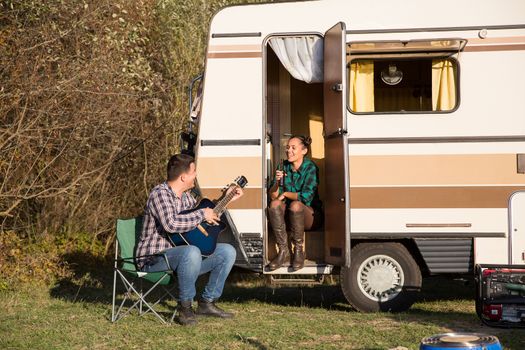 Woman enjoying a beer while her boyfriend is playing on their old guitar in front of their retro camper van.