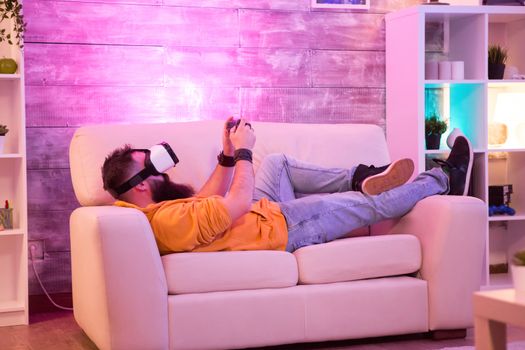 Man playing games using virtual reality goggles while sitting on sofa in a room with colorful neon light.