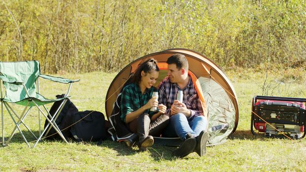 Beautiful couple having a conversation in camping tent in the wilderness and drinking hot drink. Camping essentials.