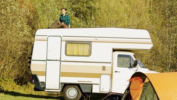 Young girl relaxing on top of retro camper van in mountain wilderness while her boyfriend going in camping tent.