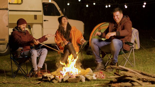 Bearded man telling a funny joke to his friends around camp fire. Retro camper van. Camping tent.