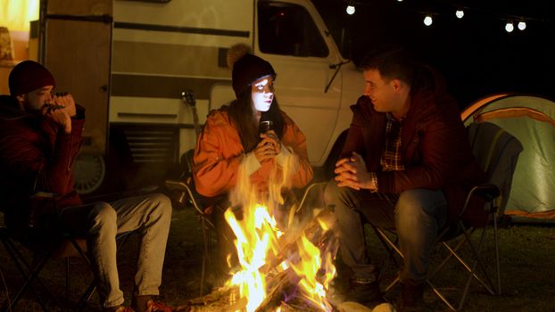 Girl telling a scary story to her friends around camp fire in cold night of autumn. Retro camper van. Light bulbs
