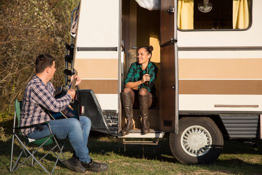 Girlfriend sitting on the stairs of retro camper van and listening her boyfriend playing on guitar.