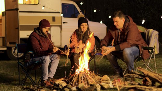 Girl warming up her hands while her friend making the fire stronger. Camp fire. Retro camper. Light bulbs.