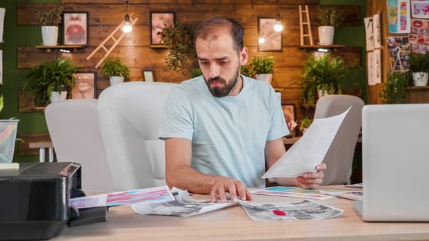 A designer boy sitting at the table and looking at some printed pictures. Creativ office