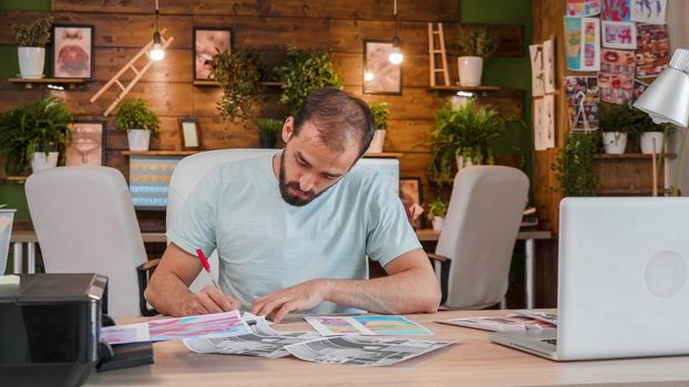 Young designer in front of some printed sketches and making notes. Modern agency office