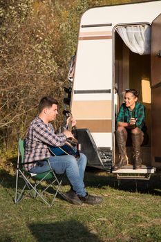 Young man trying to sing a song on guitar for his girlfriend in front of their retro camper van. Cheerful couple.