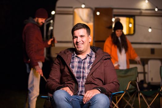 Portrait of young man smiling sitting on a camping chair with friend holding a beer in the background. Retro camper van.