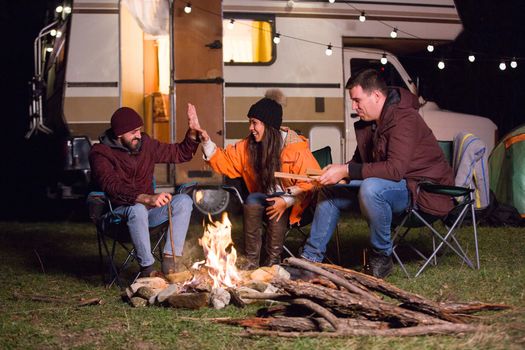 Group of close friends around camp fire laughing with retro camper van in the backgroud. Friends camping in the mountains.