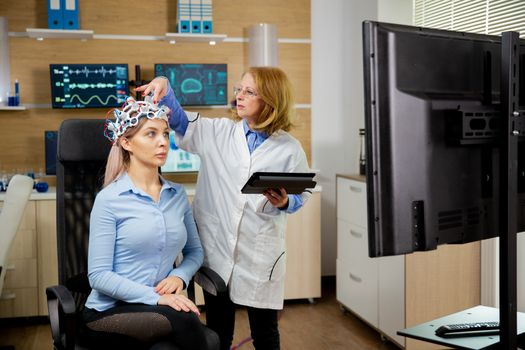 Patient woman with brain waves scanning device adjusted by doctor with tablet holding in hand. Neuroscience and brain activity