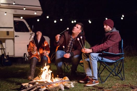 Girl laughing and holding hot marshmallow while her friends ar roasting marshmallows. Retro camper van. Camping in mountains.