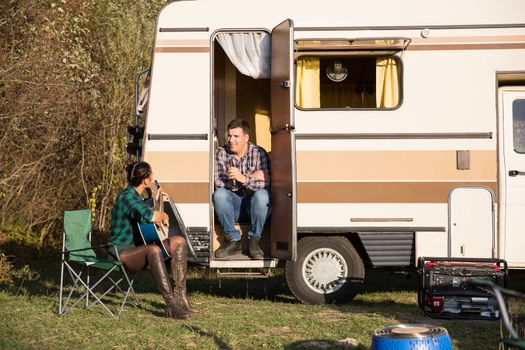 Boyfriend enjoying to listen his girlfriend singing on guitar for him in front of their retro camper. Couple relaxing in mountains.