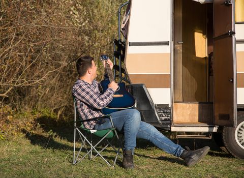 Happy hipster young man relaxing playing on guitar in front of his retro camper van in the mountains.