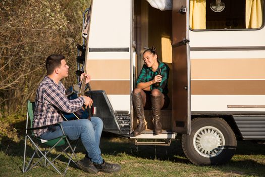 Cheerful enjoying their time together in the moutains forest with their retro camper van. Boyfriend playing a song on guitar for his girlfriend.