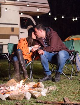 Boyfriend kissing his girlfriend near warm camp fire in a cold night of autumn in the mountains with retro camper van in the background.